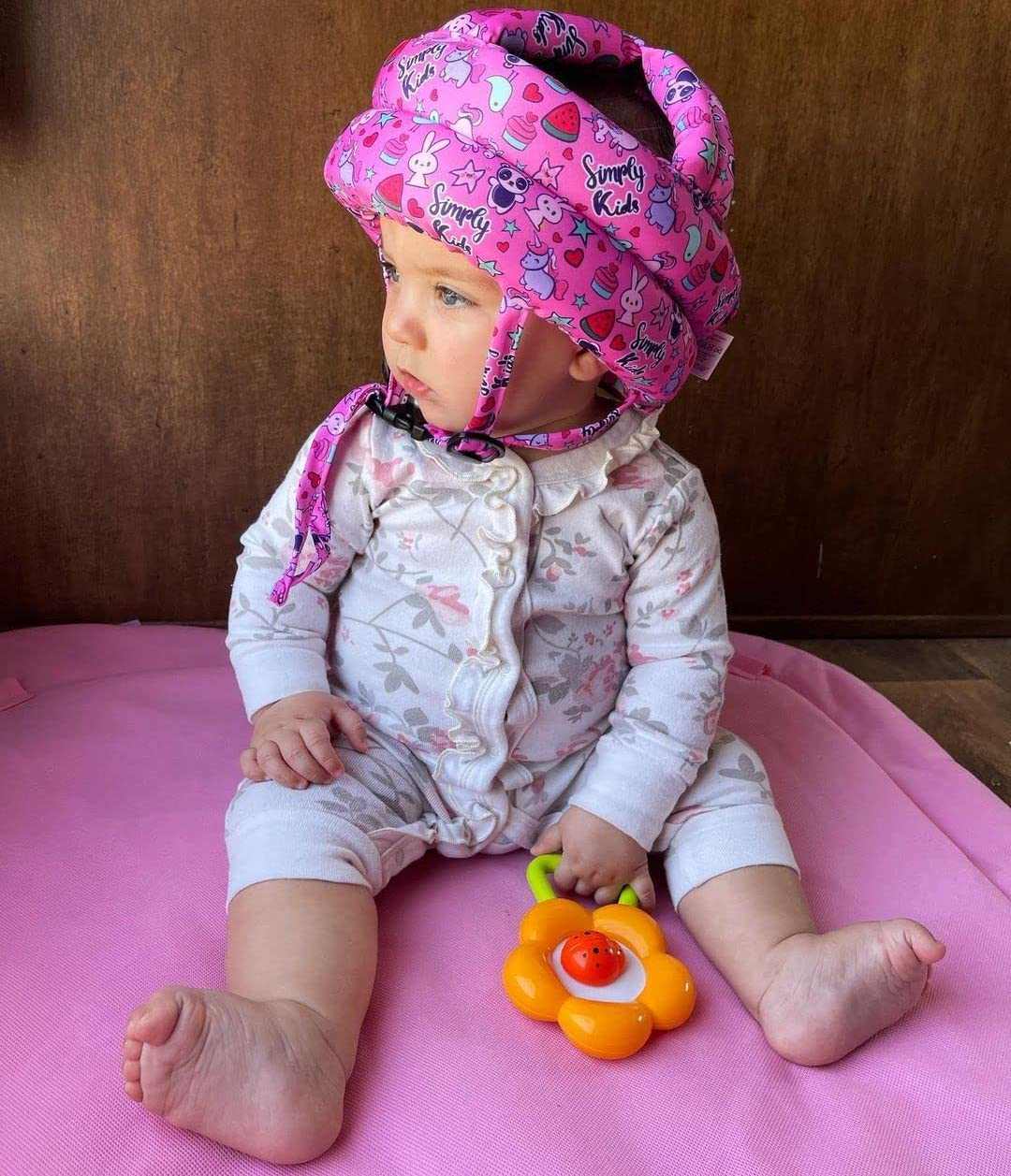 Infant wearing pink baby safety helmet with 360° head protection holding toy sitting on pink mat