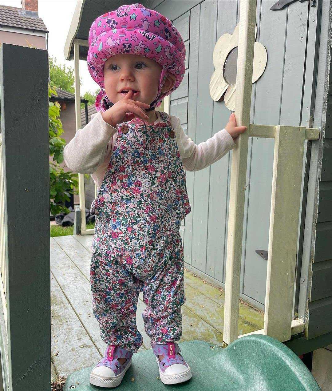 Child wearing a pink helmet and floral overalls standing in front of a small building.