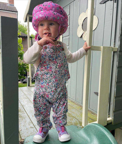 Child wearing a pink helmet and floral overalls standing in front of a small building.